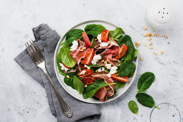 Spring Salad with spinach, cherry tomato, mozzarella, pine nuts and ham with olive oil in a simple ceramic plate on an old concrete gray background. - Fotó, kép