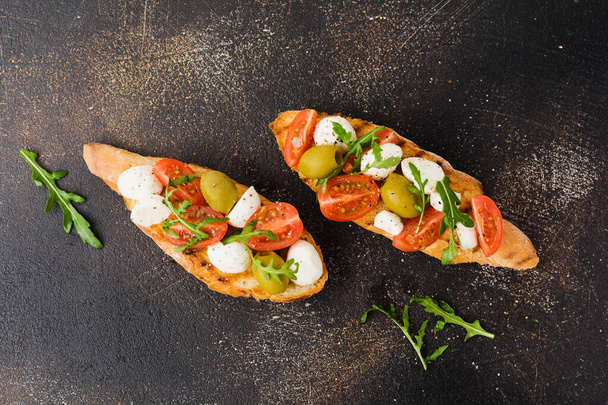 Caprese bruschetta toasts with cherry tomatoes, mozzarella, olives and basil on old dark background. Top view. - Foto, imagen