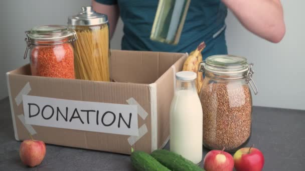 Spendenbox. Erwachsener Kaukasier füllt Karton mit Lebensmitteln auf dunklem Tisch - Filmmaterial, Video