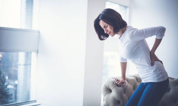 Struggling to walk. Young woman, who has just stood up from sofa, is holding her lower back while suffering from severe back pain. - 写真・画像