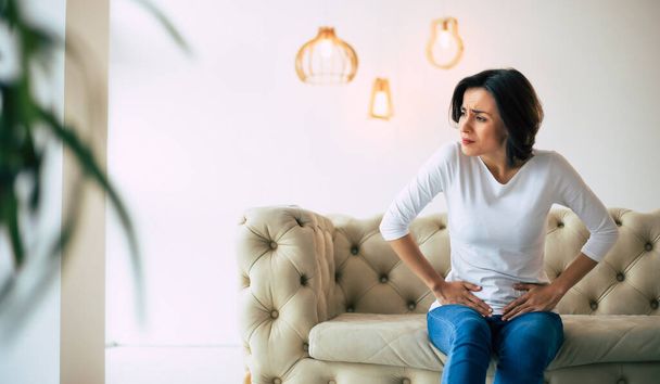 Menstrual disorder. Young woman is sitting on a sofa and touching her lower stomach while suffering from period pain. - Foto, afbeelding