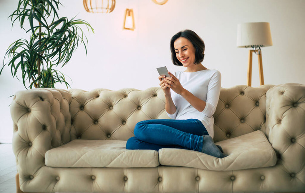 Happy face. Charming young woman is sitting on a sofa in her stylish apartment and smiling, while typing a message for someone. - Foto, immagini