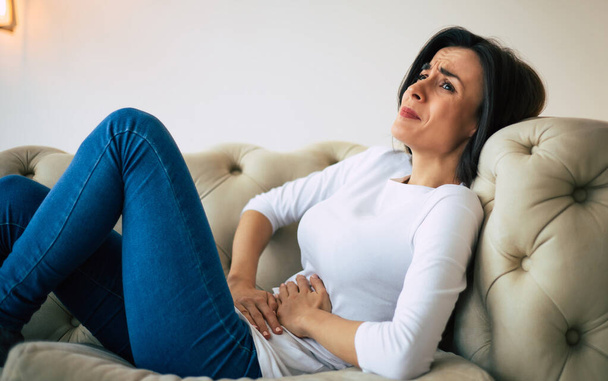 Abdominal pain. Young brunette woman is lying on a couch and crying while holding her stomach. - Photo, Image