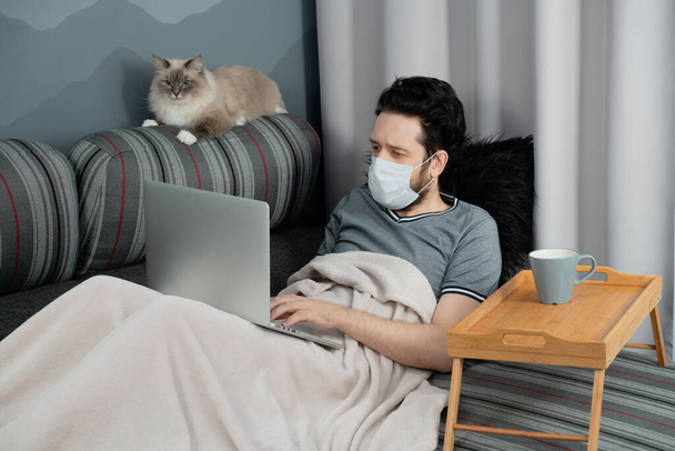 Un homme dans un masque médical travaille sur un ordinateur portable assis à la maison sur le canapé et un chat est assis à côté de lui
 - Photo, image