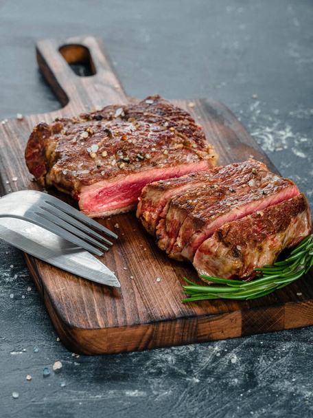 Beef steak with coarse salt and a sprig of rosemary on a wooden cutting board against a dark background - Foto, Imagem