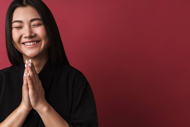 Close up de um sorriso bonito jovem asiático mulher vestindo roupas casuais de pé isolado sobre fundo vermelho, olhos fechados, implorando
 - Foto, Imagem