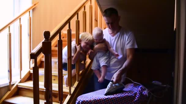 Joven padre sosteniendo bebé y camisa de planchar. Papás en casa con dos niños dedicados a las tareas domésticas
. - Metraje, vídeo