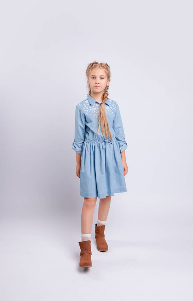 Μακριά μαλλιά κορίτσι στέκεται σε ένα ανοιχτό γκρι φόντο σε ένα μπλε φόρεμα. - Φωτογραφία, εικόνα