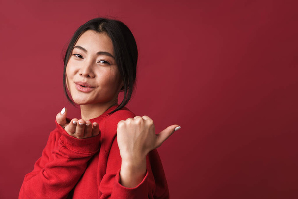 Close up of a pretty smiling young asian woman wearing casual clothes standing isolated over red background, pointing finger away at copy space, sending kiss - Foto, Imagem