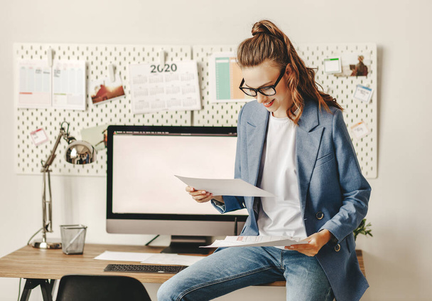 Молода жінка-підприємець в повсякденному вбранні сидить на столі і читає документи, задоволені великими результатами під час роботи в сучасному робочому місці
 - Фото, зображення