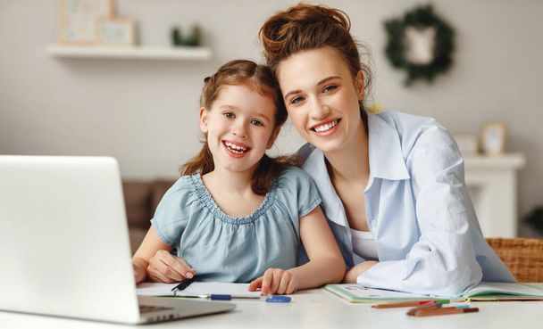 Позитивная молодая женщина помогает дочери в поиске информации для домашней работы в Интернете, сидя вместе за столом с ноутбуком в доме
 - Фото, изображение