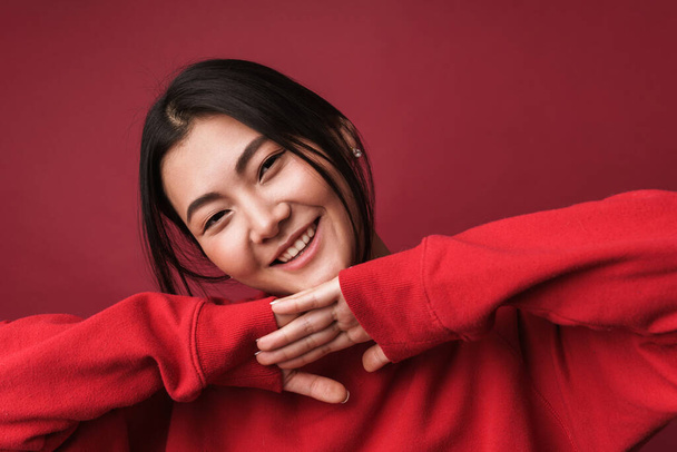 Close up of a pretty smiling young asian woman wearing casual clothes standing isolated over red background, holding arms at face - Photo, Image