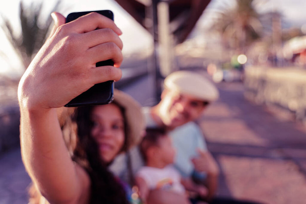 Szczęśliwa rodzina w parku robi selfie w słoneczny dzień. Abstrakcyjna rozmyta niezidentyfikowana osoba używa aparatu selfie. Zamazany obraz rodziny siedzącej na zewnątrz i pozującej do selfie. Zdjęcie.. - Zdjęcie, obraz