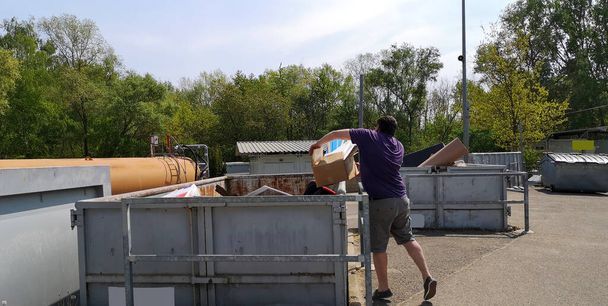 Ein dicker, dunkelhaariger Mann mittleren Alters wirft an einem sonnigen Tag in Deutschland, Europa Pappkartons mit Hausmüll in einen riesigen Container an der Sammelstelle für Großmüll.. - Foto, Bild