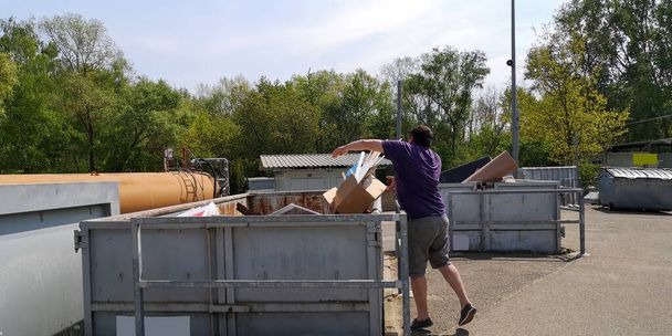 Толстый, темноволосый мужчина средних лет бросает картонную коробку с бытовыми отходами в огромный контейнер на станции сбора крупногабаритного мусора в Германии, Европе в солнечный день.
. - Фото, изображение