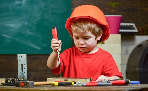 Kid boy in orange hard hat or helmet, study room background. Little helper concept. Boy play as builder or repairer, work with tools. Child dreaming about future career in architecture or building. - Foto, Bild