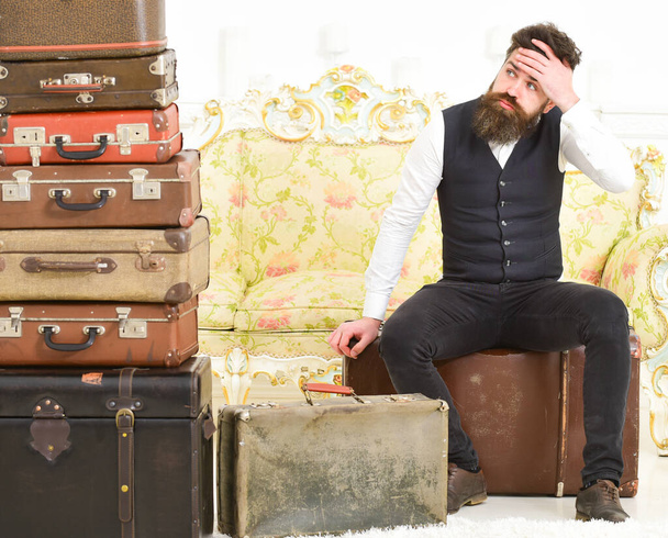 Macho elegant on tired face sits, exhausted at end of packing, near pile of vintage suitcases. Man with beard and mustache packed luggage, white interior background. Luggage and relocation concept. - Foto, Imagem