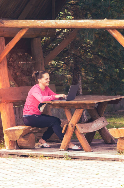 Laughing cheerful young girl working on a laptop in a wooden arbor on the nature. - Photo, Image