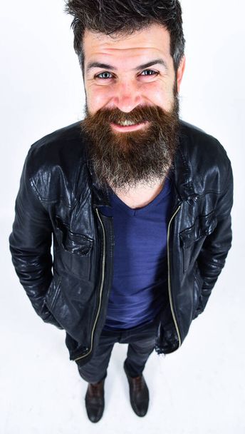 Man with beard and mustache on smiling face looking at camera. Macho wears leather jacket, white background. Hipster looks cheerful while posing in stylish outfit. Menswear and fashion concept. - 写真・画像