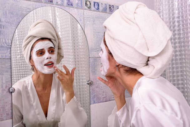 Hermosa mujer morena en casa en pijama y toalla en su cabeza puso máscara blanca en su cara en el baño
 - Foto, Imagen