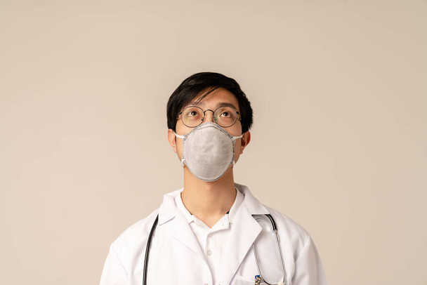 Изображение молодого врача-азиата в белой медицинской форме и защитной маске, стоящей изолированно на бежевом фоне
 - Фото, изображение