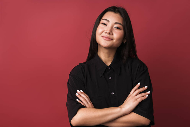 Close up of a pretty smiling young asian woman wearing casual clothes standing isolated over red background, arms folded - Fotografie, Obrázek