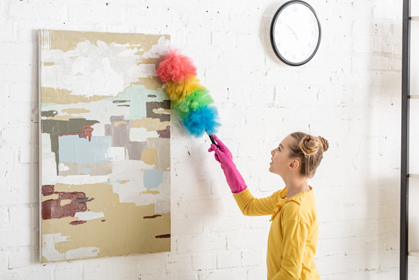 Cute child in rubber gloves sweeping away dust from picture with feather duster in living room - Photo, Image