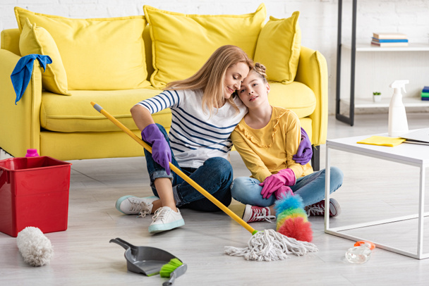 Tired mother hugging daughter with cleaning supplies on floor near coffee table in living room - Foto, imagen