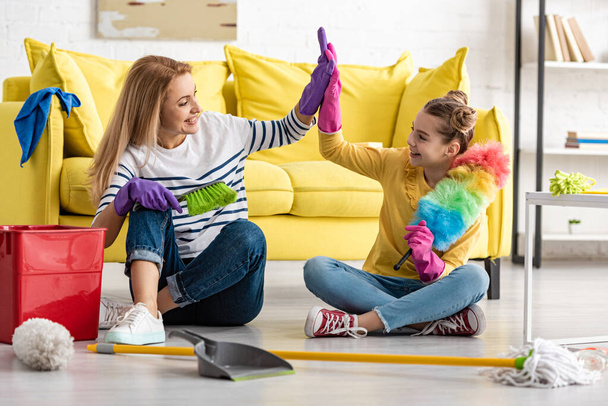 Mother and cute daughter with cleaning supplies giving high five, smiling and looking at each other on floor in living room - Foto, afbeelding