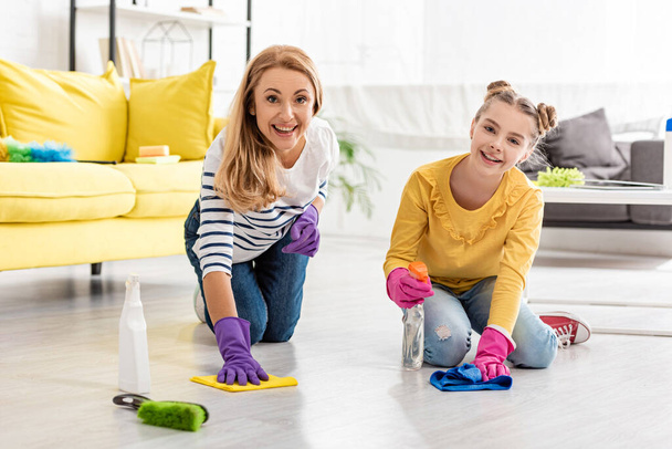 Mother and daughter with spray bottle and rags wiping floor, smiling and looking at camera near brush in living room - Foto, immagini