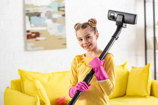 Cute child with vacuum cleaner smiling and looking at camera in living room - Φωτογραφία, εικόνα