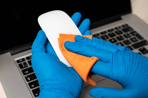 Καθαρισμός και απολύμανση ποντικιού με προστατευτικά γάντια. Καθαρισμός επιφανειών φορητού υπολογιστή από ιούς, μικρόβια και βρωμιά στο γραφείο για ασφαλή εργασία. Έννοια πρόληψης του ιού του κερατοειδούς.  - Φωτογραφία, εικόνα