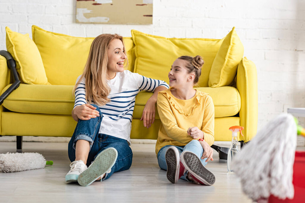 Selective focus of mother and cute daughter looking at each other and smiling near sofa and cleaning supplies on floor in living room - Photo, Image