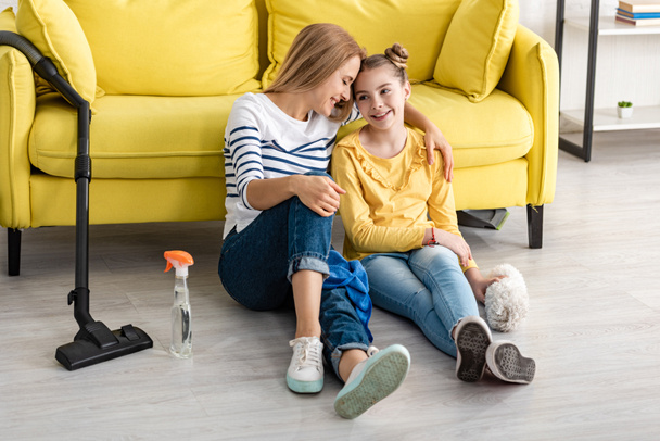 Madre abrazando linda hija y sonriendo cerca de sofá y artículos de limpieza en el suelo en la sala de estar
 - Foto, Imagen