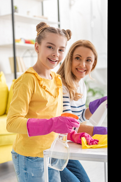 Cute child holding rag and spray bottle, smiling and looking at camera with mother near coffee table in living room - Foto, Imagem