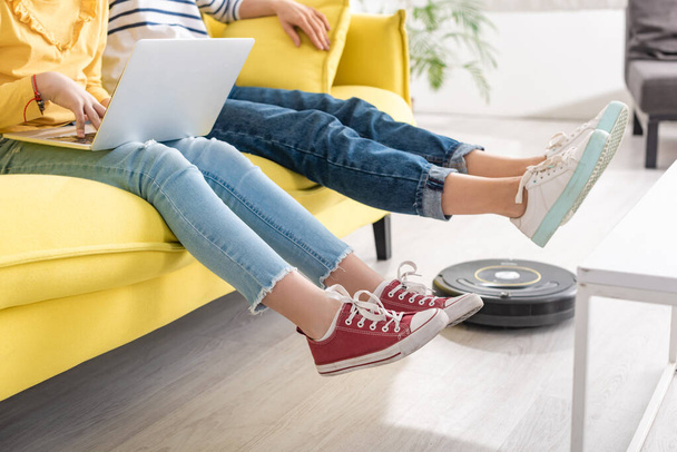 Vista ritagliata di figlia con laptop vicino alla madre con gambe sollevate sul divano e aspirapolvere robotico sul pavimento in soggiorno
 - Foto, immagini