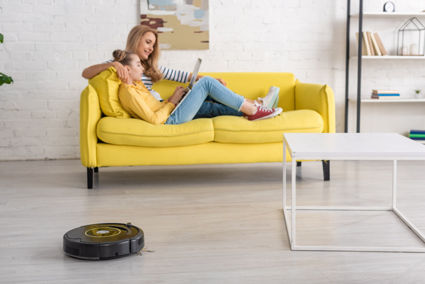 Mother and daughter with laptop resting on sofa near coffee table and robotic vacuum cleaner on floor in living room - Φωτογραφία, εικόνα