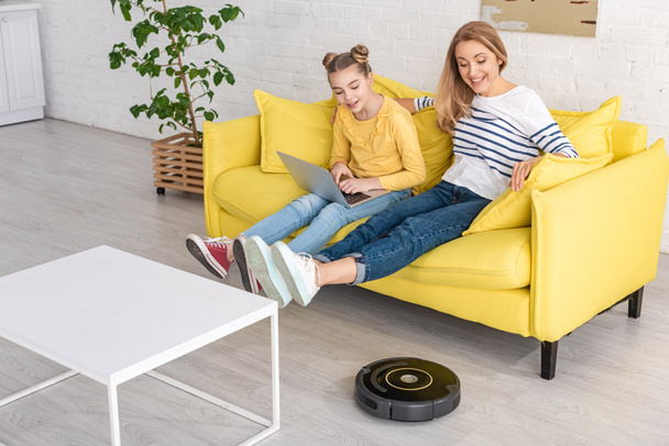 Cute child with laptop near mother with raised legs looking at robotic vacuum cleaner near coffee table in living room - Photo, Image