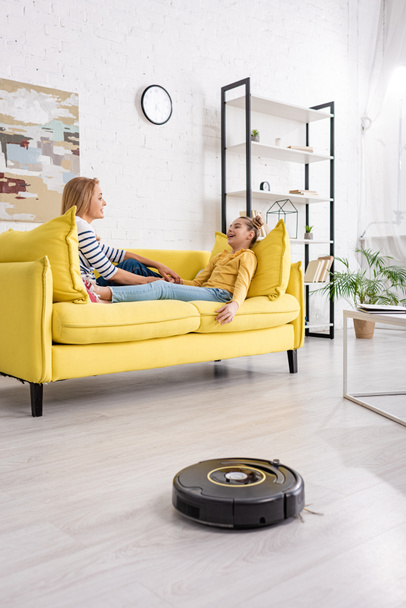 Mother and daughter looking at each other and lying on sofa near coffee table and robotic vacuum cleaner on floor in living room - Foto, Bild