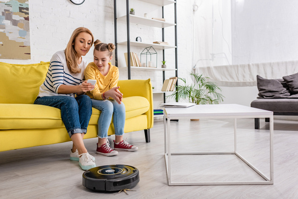 Мать и дочь смотрят на смартфон на диване возле журнального столика с ноутбуком и роботизированным пылесосом на полу в гостиной
 - Фото, изображение