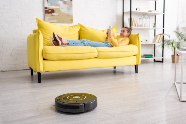 Selective focus of kid with smartphone lying on sofa near robotic vacuum cleaner on floor in living room - 写真・画像