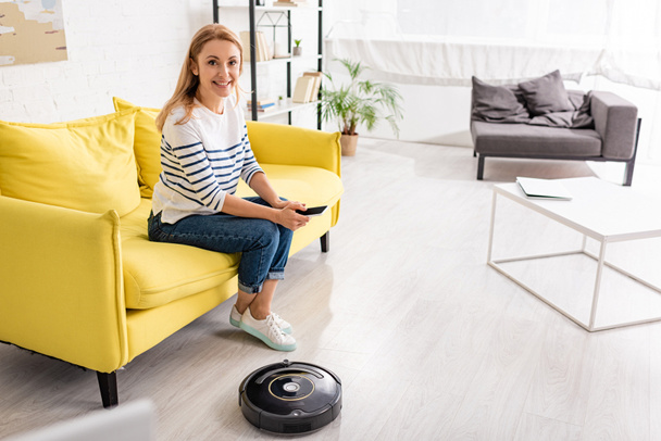 Beautiful woman with smartphone smiling and looking at camera on sofa near coffee table and robotic vacuum cleaner on floor in living room - Foto, afbeelding