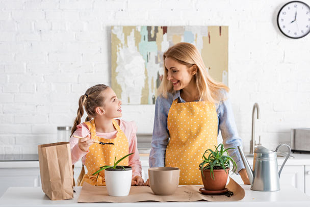 Mother and daughter looking at each other and smiling near table with watering pot, paper bag and flowerpots in kitchen - Fotoğraf, Görsel