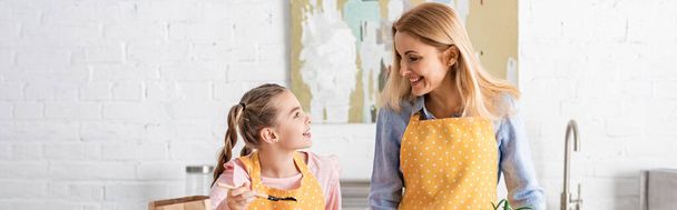 Panoramic orientation of mother and daughter looking at each other and smiling in kitchen - Photo, Image