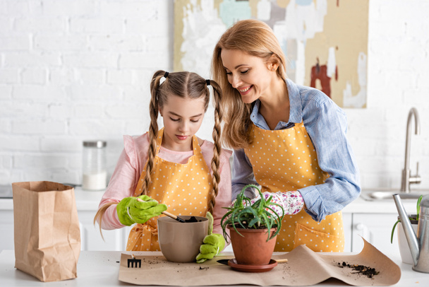 Frau lächelt neben süßer Tochter und hält Schaufel mit Erde in Blumentopf neben Tisch in Küche - Foto, Bild