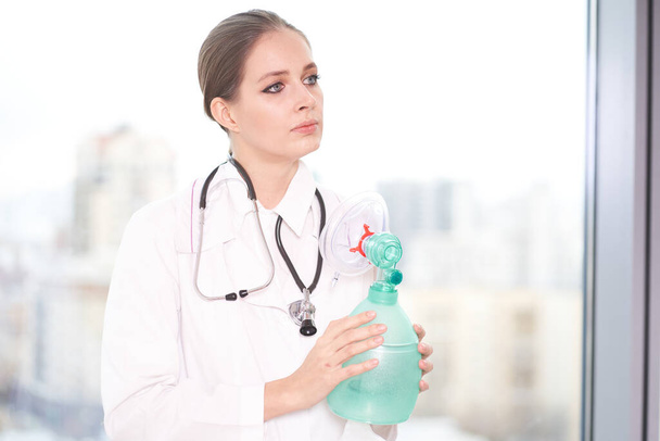   Een jonge vrouwelijke arts staat in een witte jas, kijkt weg, houdt een ventilator vast. Gezondheidszorg, quarantaine, pandemie                              - Foto, afbeelding
