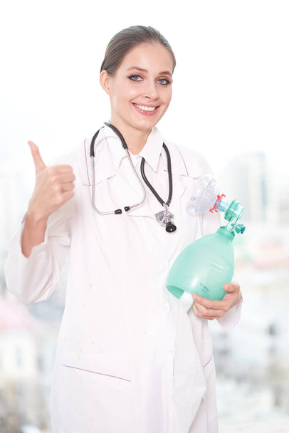 Retrato de una joven doctora, sosteniendo un respirador con la mano, mostrando la clase con la otra mano, sobre un fondo blanco, mira a la cámara y sonríe
                                  - Foto, Imagen