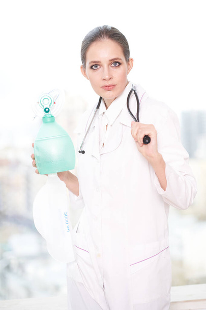 Retrato de uma jovem médica segurando um estetoscópio com a mão e um ventilador com a outra mão, em um fundo branco, ela olha para a câmera e sorri
                                 - Foto, Imagem