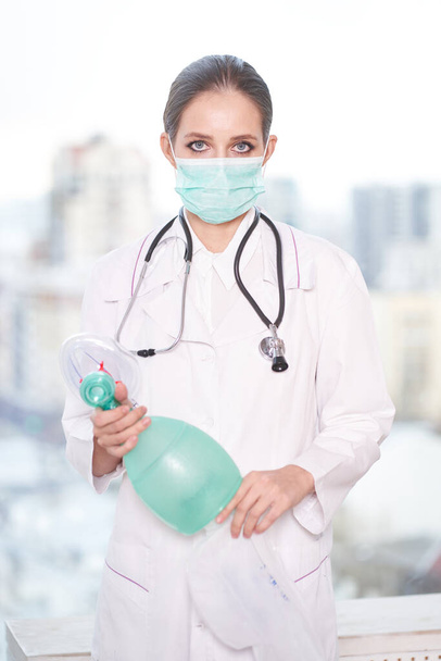 Μια νεαρή γυναίκα γιατρός στέκεται σε ένα λευκό παλτό, φορώντας μια μάσκα μιας χρήσης, κρατώντας έναν αναπνευστήρα. Υγεία, καραντίνα, πανδημία                                  - Φωτογραφία, εικόνα
