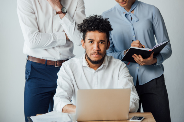 Enfoque selectivo del hombre de negocios afroamericano mirando a la cámara cerca de gente de negocios con portátil en la oficina
 - Foto, Imagen